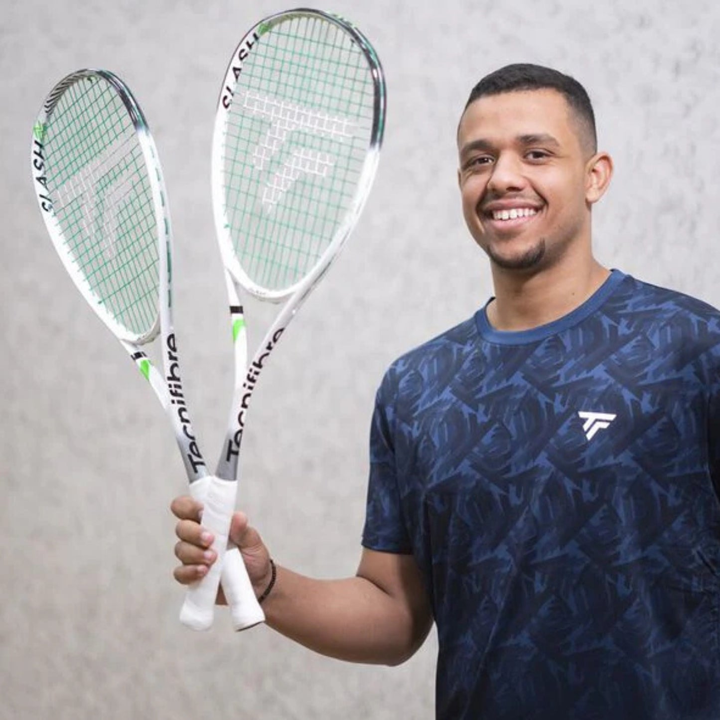 Man holding two Tecnifibre squash rackets against a gray background