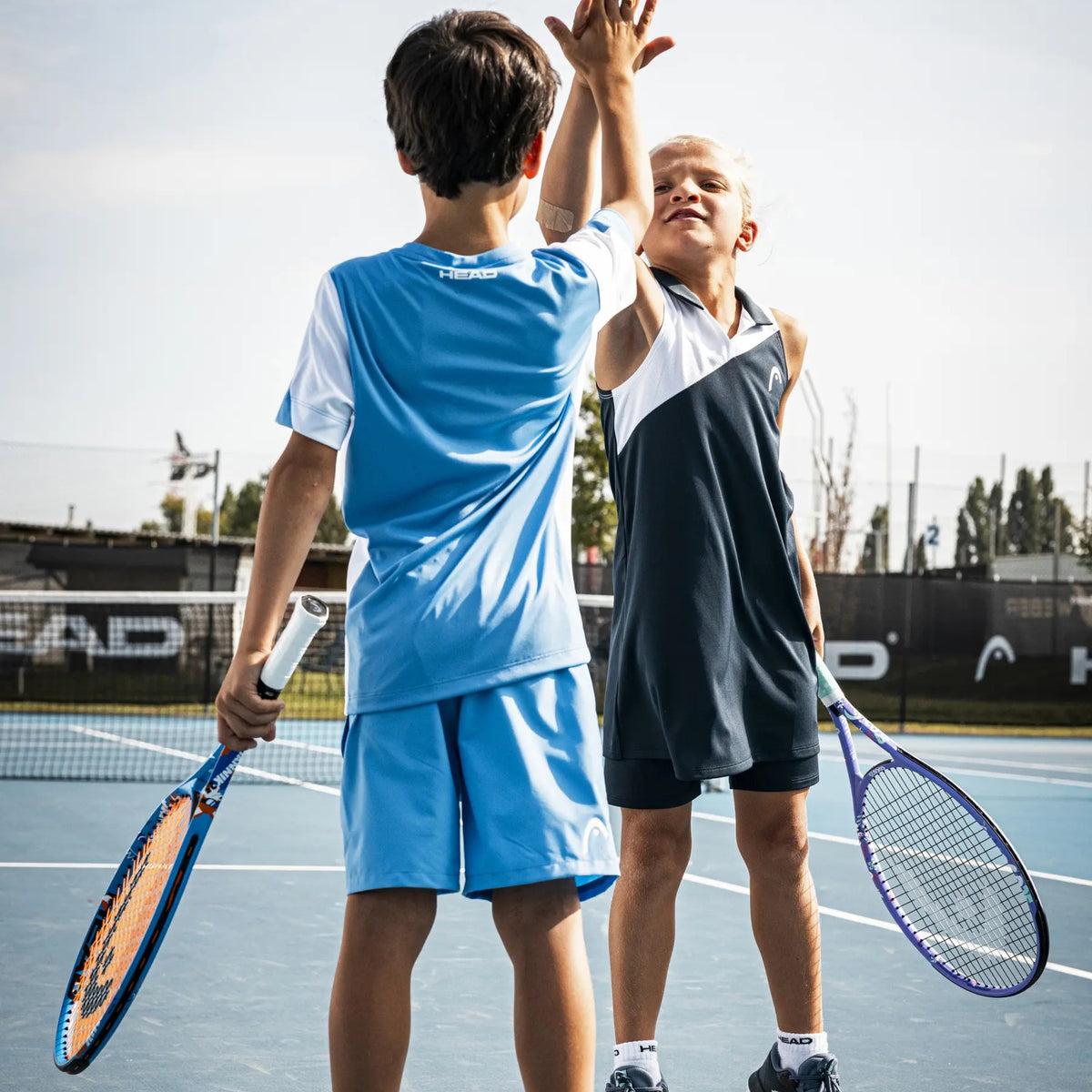 Two children playing tennis on an outdoor court, wearing athletic clothing from different brands and holding tennis rackets.