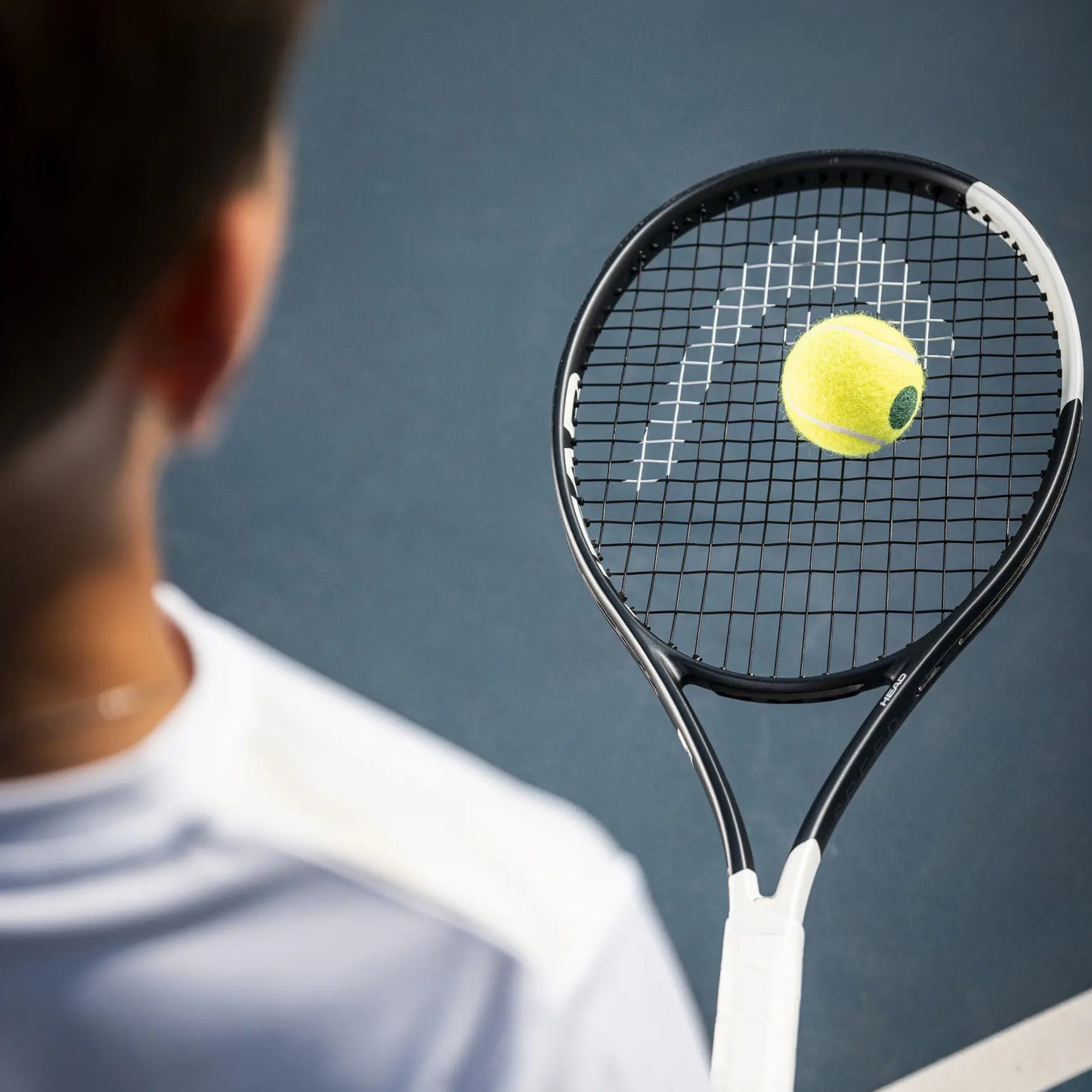 A tennis racket with a yellow tennis ball on its strings, shown from behind a blurred player on a blue tennis court.