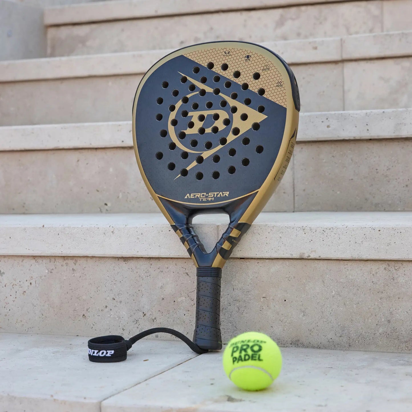 A gold and black Dunlop racket standing against the stairs with a padel ball in the picture