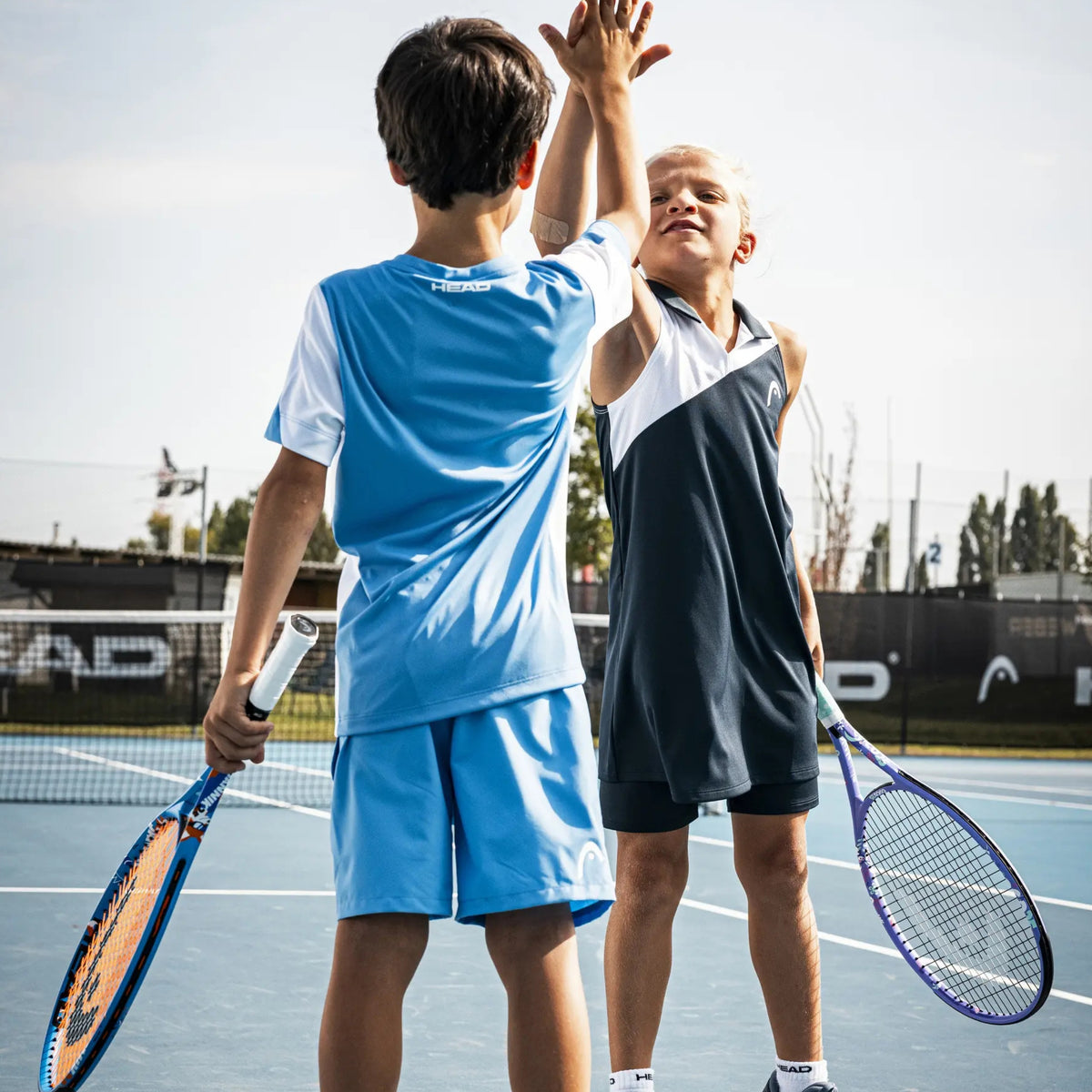 Two boys on a Head tennis court high-fiving, wearing athletic wear. One boy wears a light blue and white tennis set, and the other wears a dark navy and white sleeveless set.