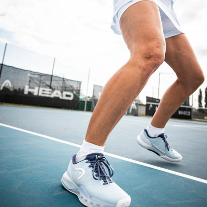 Person wearing white tennis shoes with blue laces on a tennis court, featuring HEAD branding.