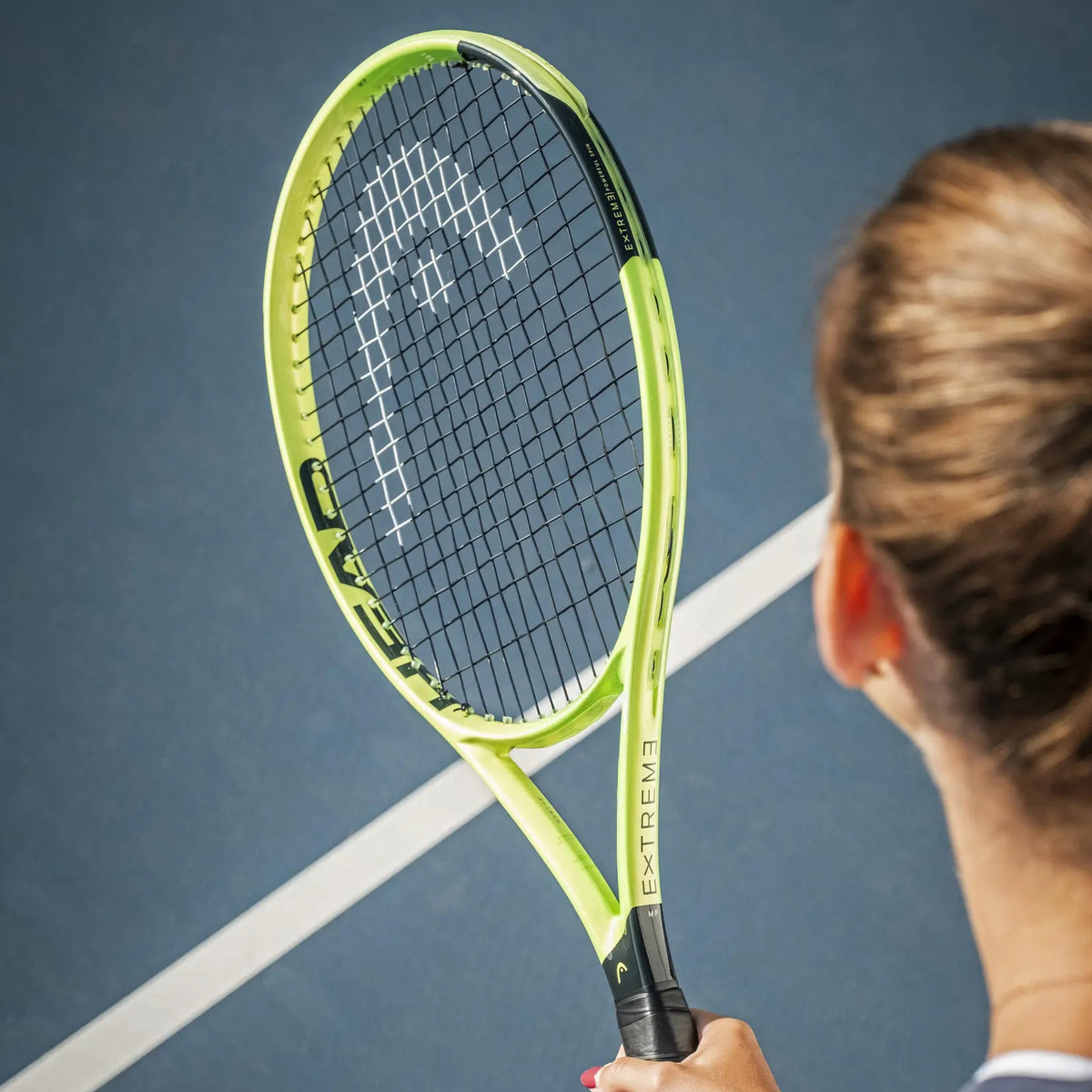 A Head tennis racket with a bright lime green and black frame, held by a player on a blue tennis court. The player is shown from behind, and the racket features the Wilson brand logo on the throat.