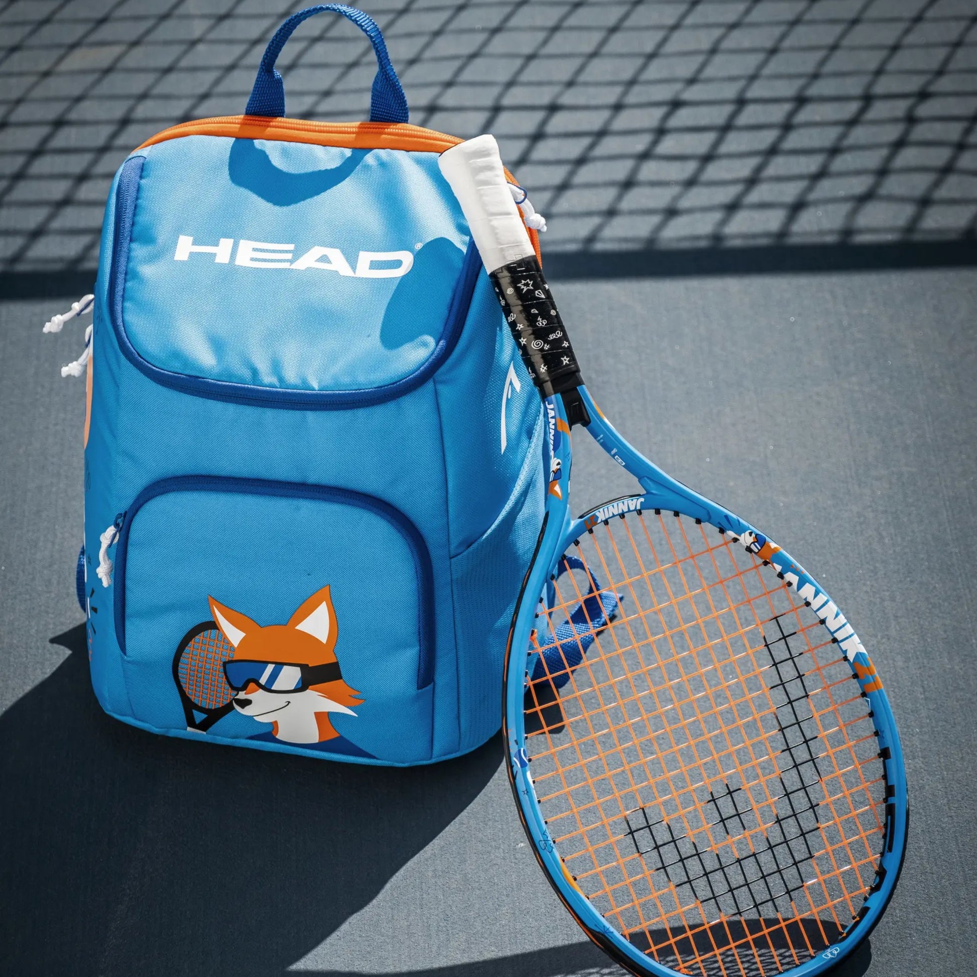 A blue HEAD tennis bag with orange accents and a cartoon fox graphic, shown next to a blue and orange tennis racket on a tennis court.