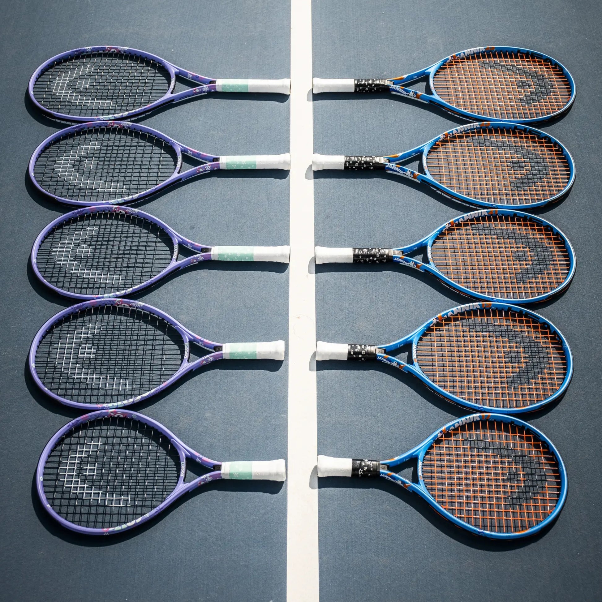 Eight Head tennis rackets arranged in two vertical rows on a blue tennis court, separated by a white center line. The rackets have purple, blue, and black frames with white and green grip tape, and brown stringing.