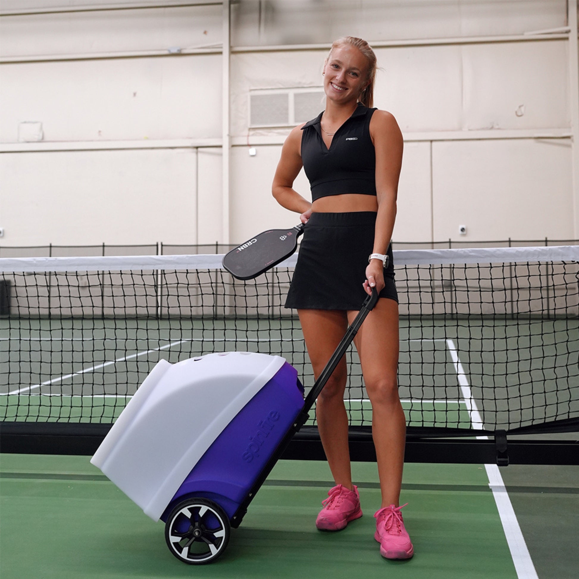 Woman on a tennis court holding a paddle and a large ball machine standing next to a net.