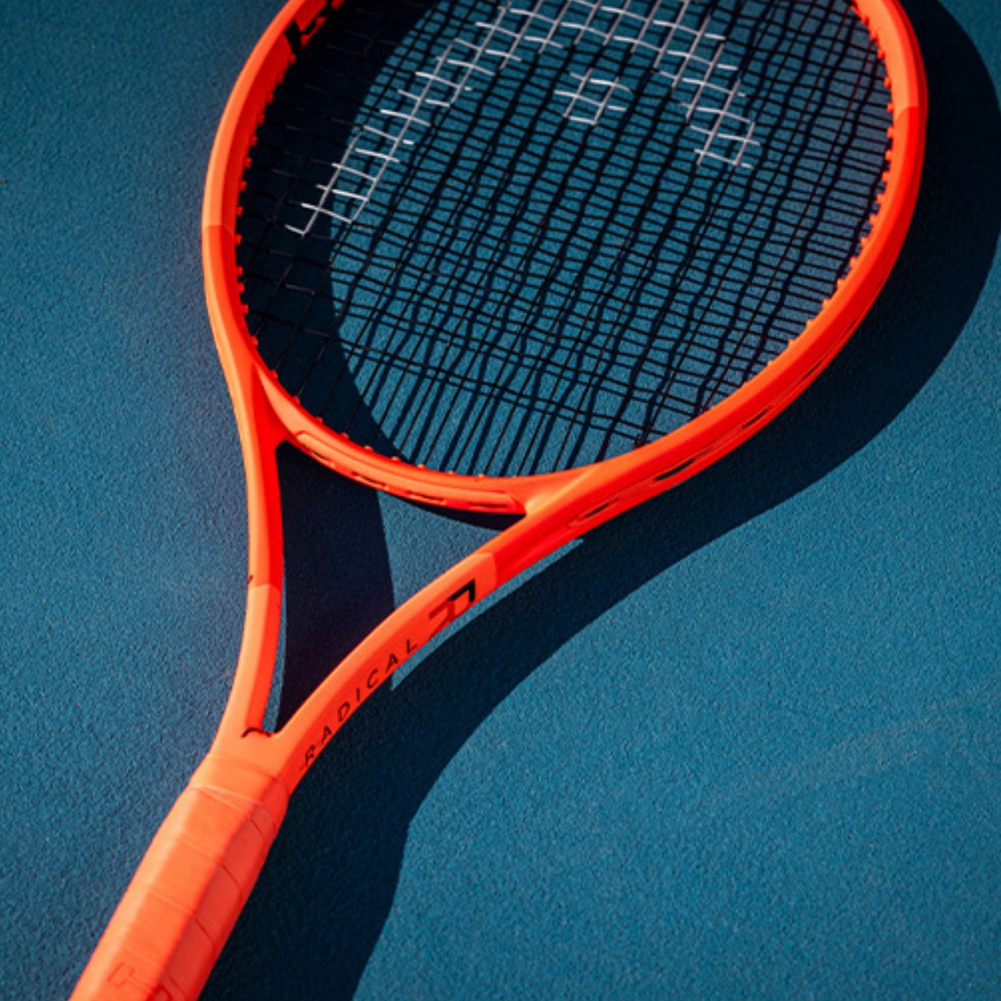 A close-up of a bright orange Head tennis racket with black strings, resting on a textured blue court surface.