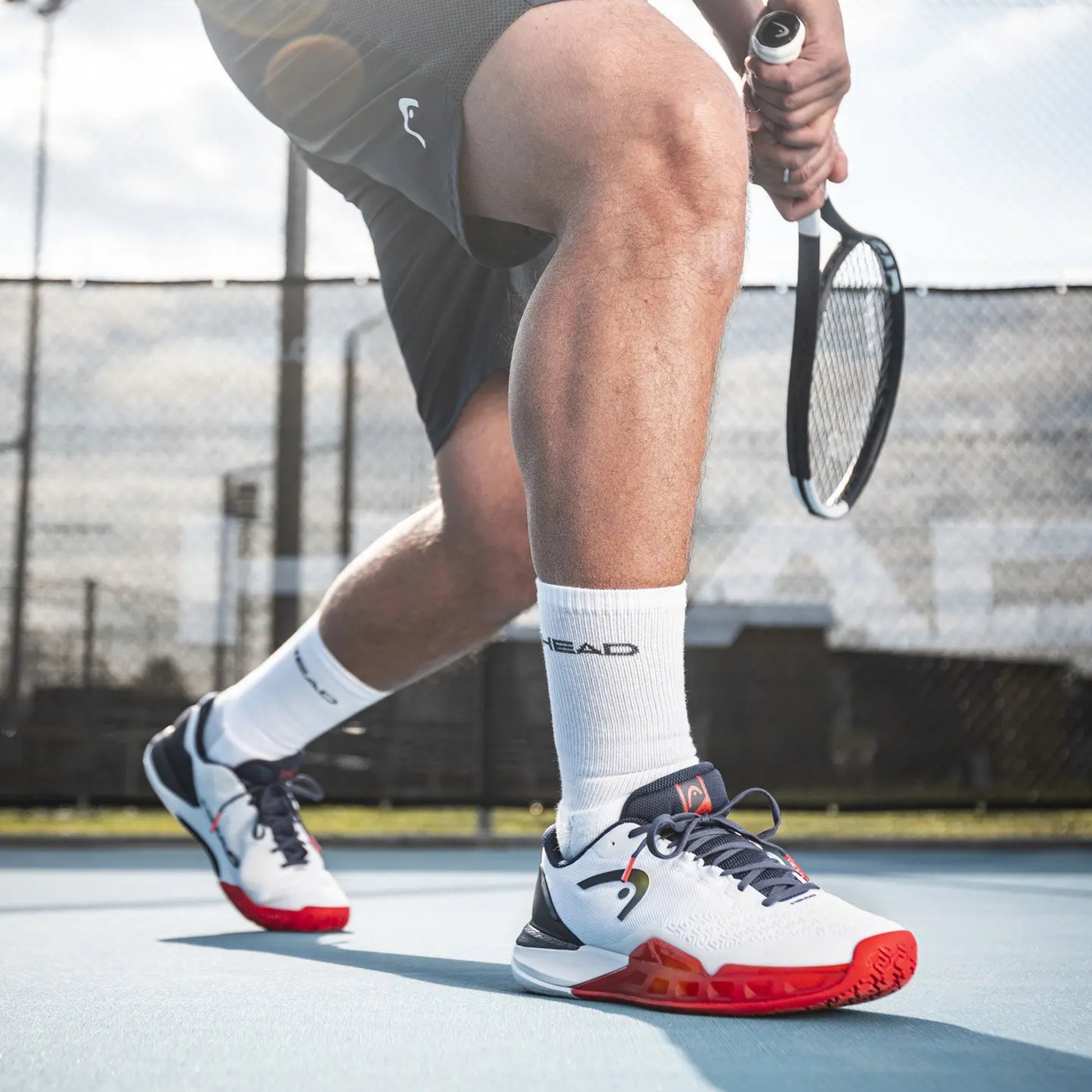 Person playing tennis on a court wearing white and red shoes with visible 'HEAD' branding.