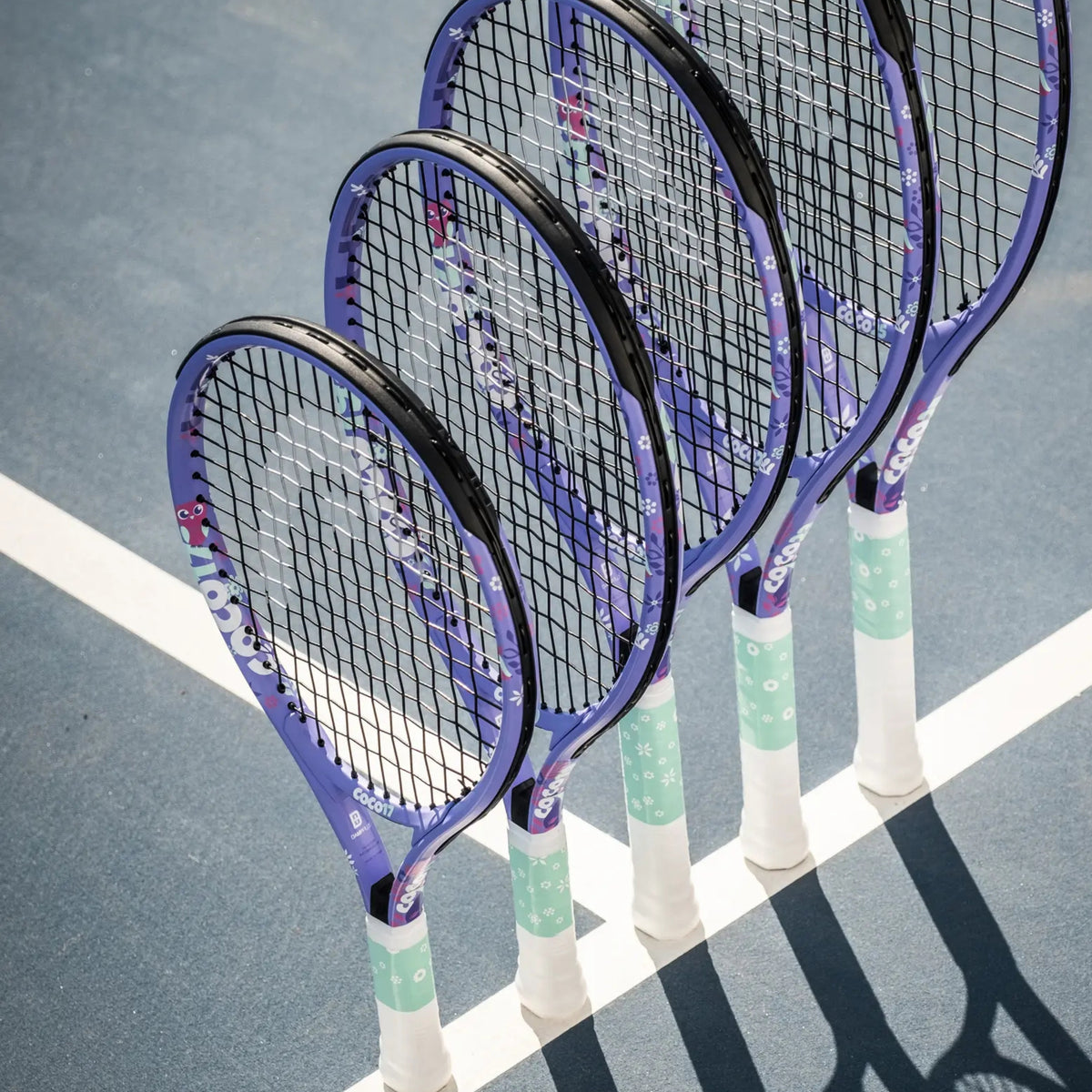 Four purple and white Head tennis rackets with black strings are lined up on a blue tennis court next to a white boundary line.