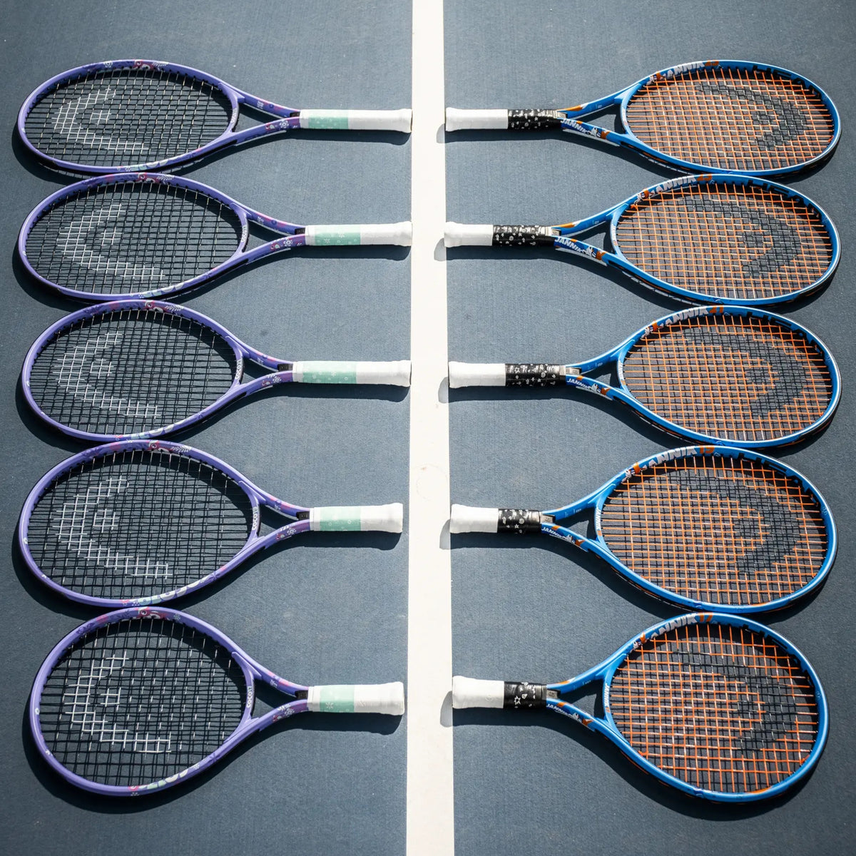 Eight Head tennis rackets arranged in two vertical rows on a blue tennis court, separated by a white center line. The rackets have purple, blue, and black frames with white and green grip tape, and the strings are orange, black, or white.