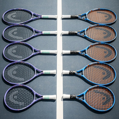 Eight Head tennis rackets arranged in two vertical rows on a blue tennis court, separated by a white center line. The rackets have purple, blue, and black frames with white and green grip tape, and the strings are orange, black, or white.