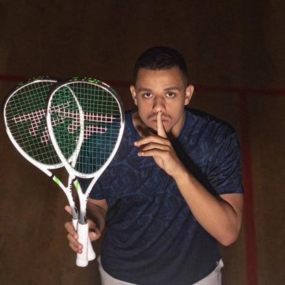 A man in a dark patterned t-shirt holding two white Tecnifibre squash rackets with green strings, posing with his finger on his lips in an indoor court setting.