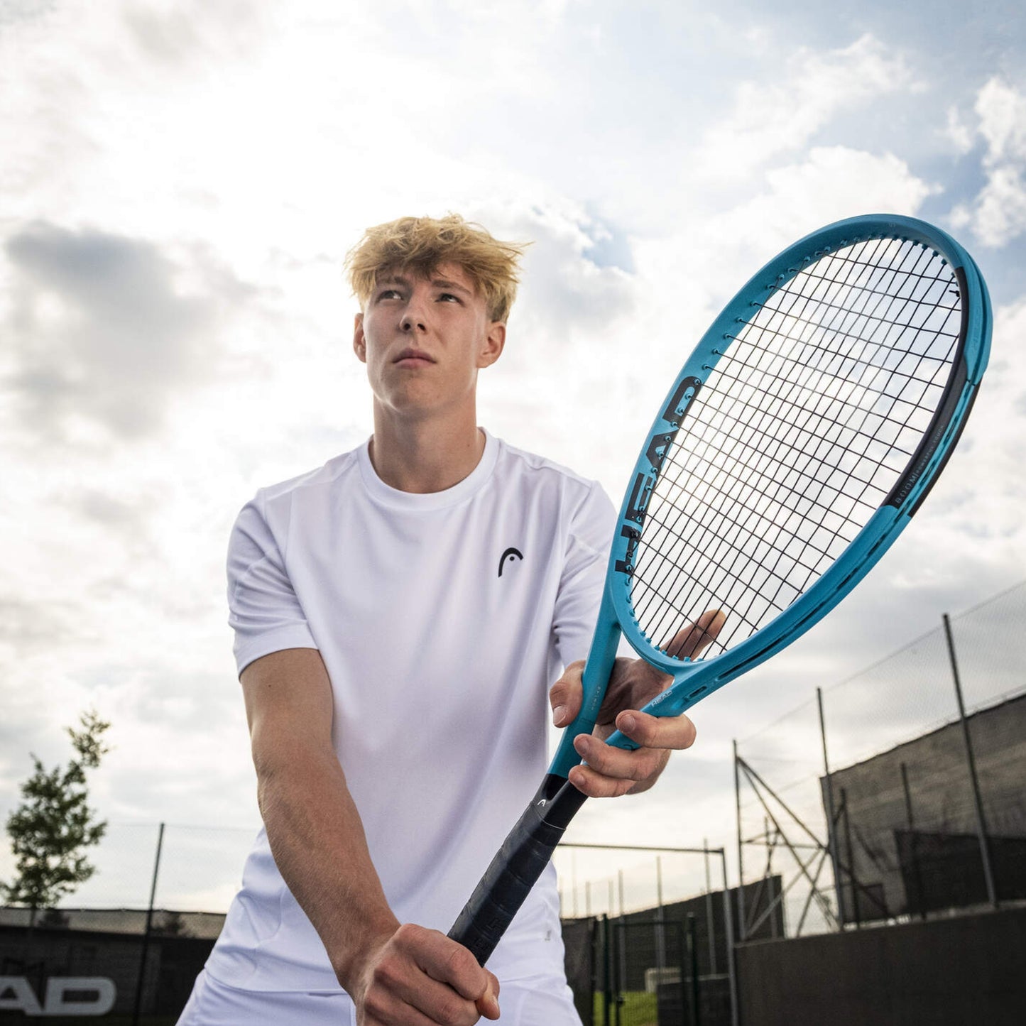 Person holding a blue Head tennis racket on a tennis court with a cloudy sky.