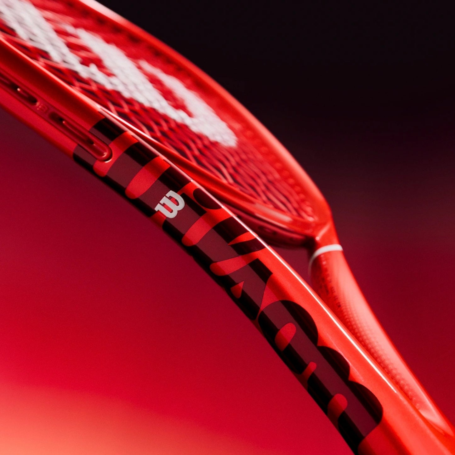 Close-up of a red and black tennis racket with white strings, featuring the Wilson logo on the frame, set against a dark red gradient background.