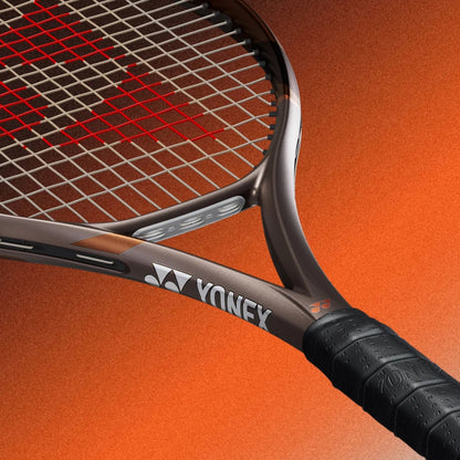 Close-up of a Yonex tennis racket showing the dark gray frame, red strings, and black grip against a textured orange background.