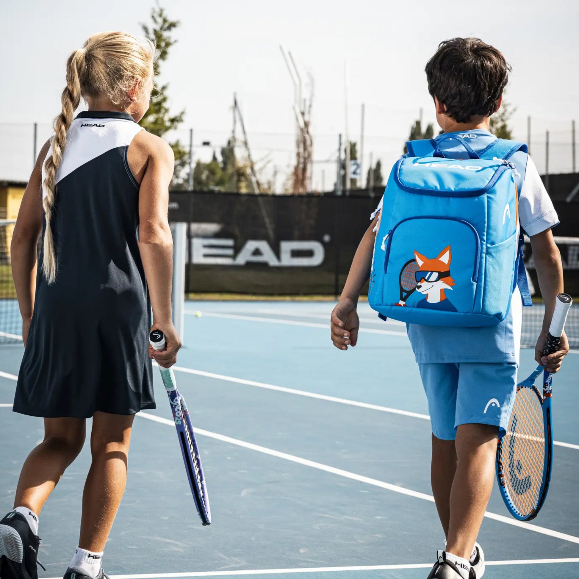 Two children on a blue tennis court, seen from behind. The girl wears a dark navy tennis dress and holds a racket, while the boy wears a light blue tennis outfit and carries a bright blue backpack with a cartoon fox design.