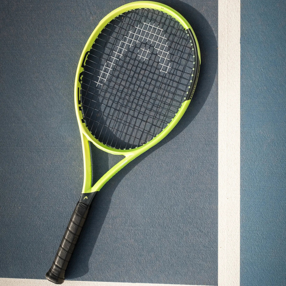 A Head tennis racket with a bright neon yellow-green frame and black grip, resting diagonally on a blue tennis court with a white boundary line.