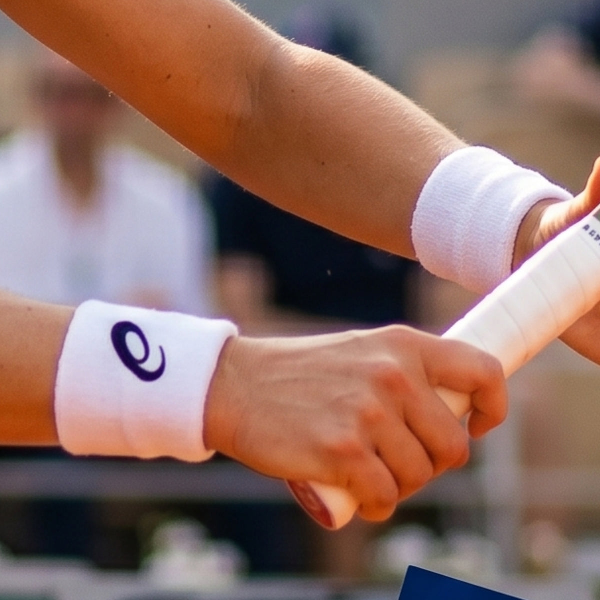 Two hands holding a tennis racket with white wristbands featuring a logo, blurred background