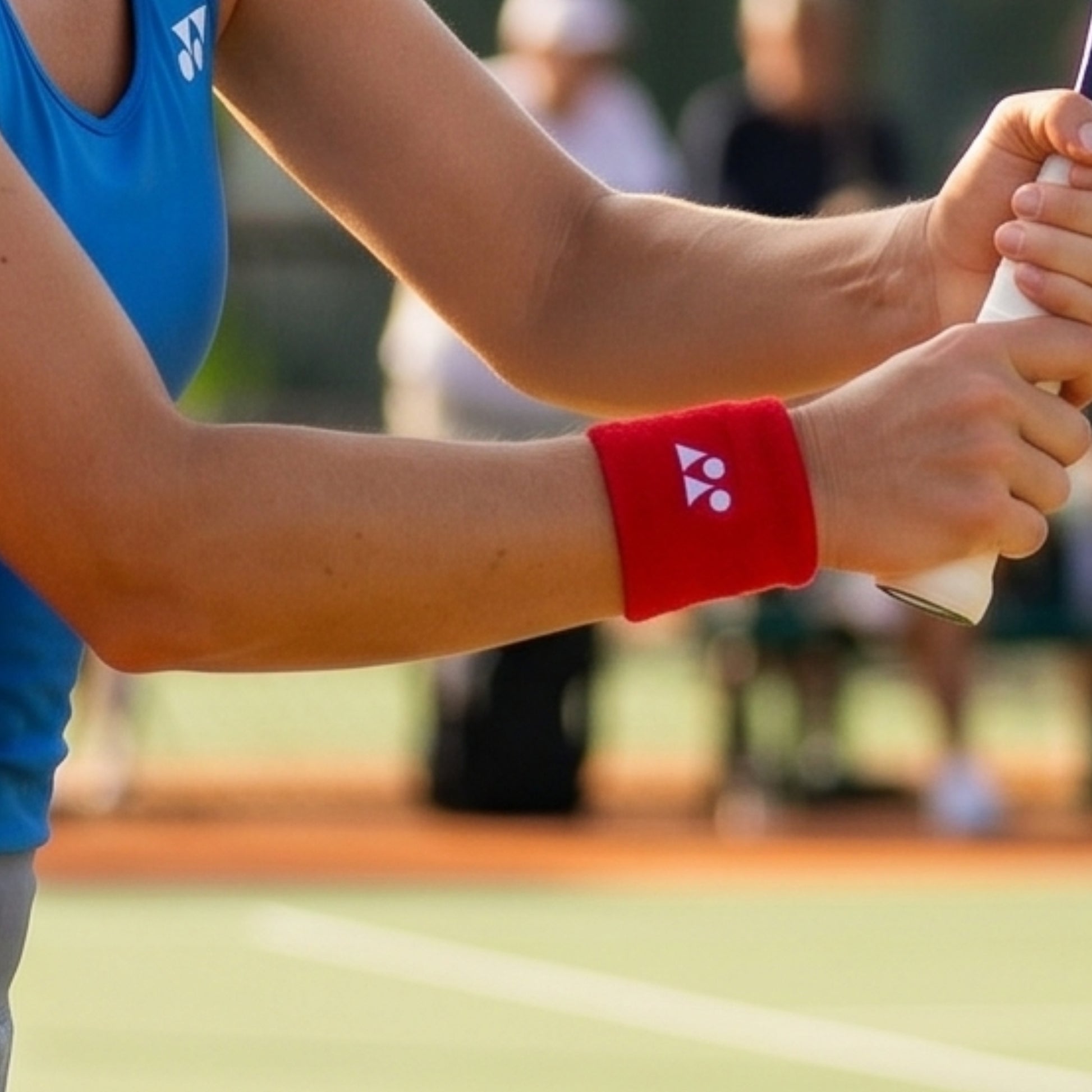 Person holding a tennis racket with a red wristband featuring a logo on a blurred tennis court background