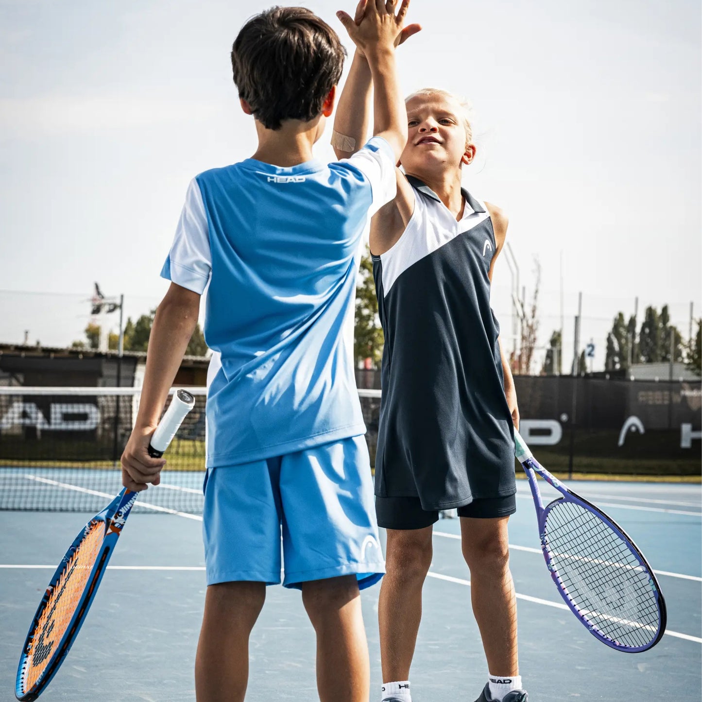 Two children on an outdoor tennis court wearing athletic apparel, one in a light blue set and one in a dark navy-and-white sleeveless set, holding tennis rackets and high-fiving at the net.