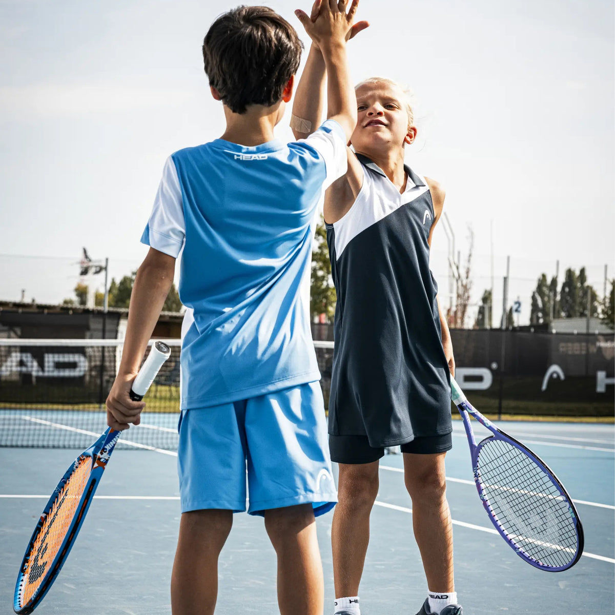 Two children on an outdoor tennis court wearing athletic apparel, one in a light blue set and one in a dark navy-and-white sleeveless set, holding tennis rackets and high-fiving at the net.