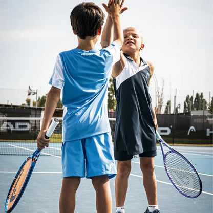 Two children on an outdoor tennis court wearing athletic apparel, one in a light blue set and one in a dark navy-and-white sleeveless set, holding tennis rackets and high-fiving at the net.