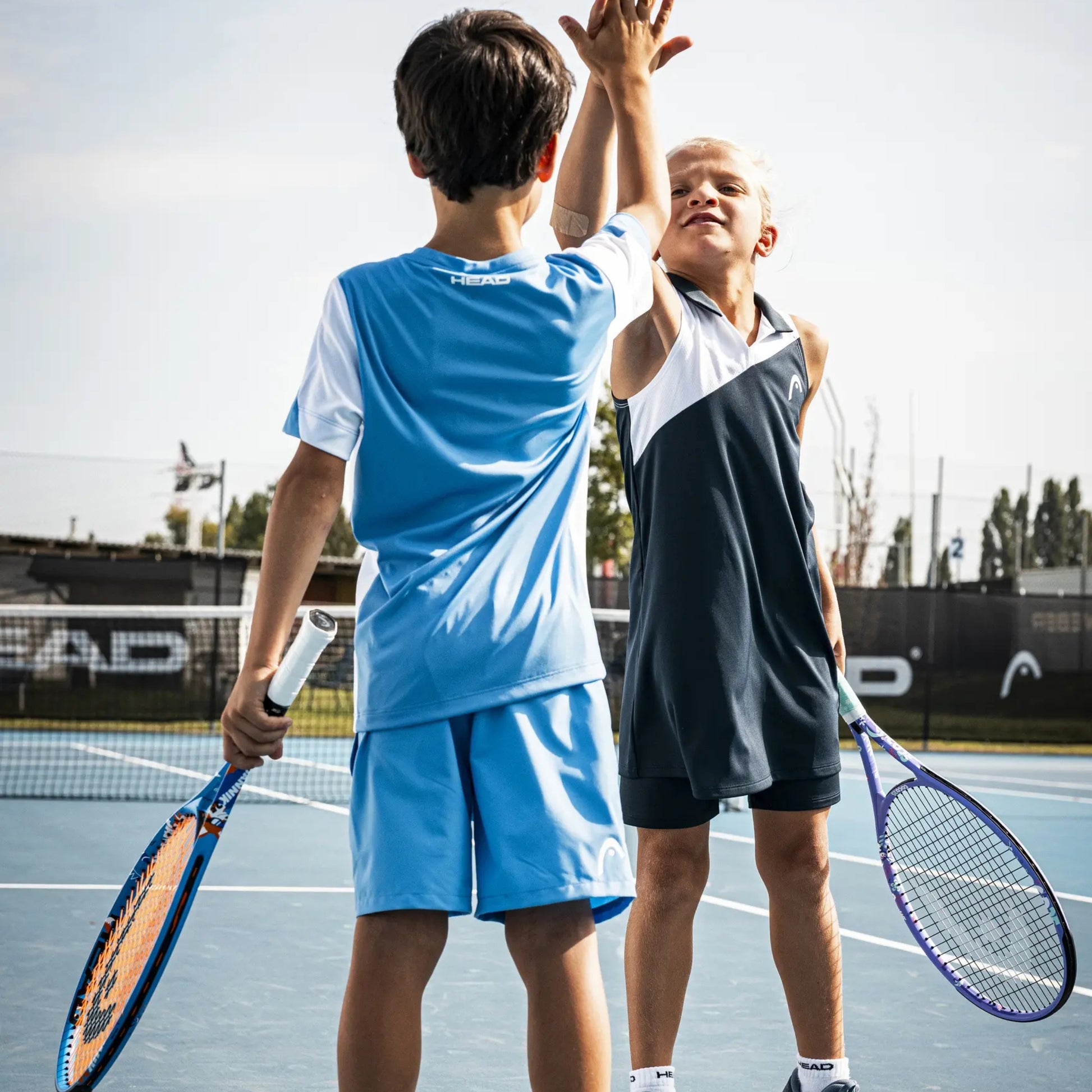 Two children playing tennis on an outdoor court, wearing athletic outfits from Adidas. One child is seen from behind in a light blue and white Adidas set, holding a blue racket, while the other child in a dark navy and white Adidas set faces him, holding a purple racket.